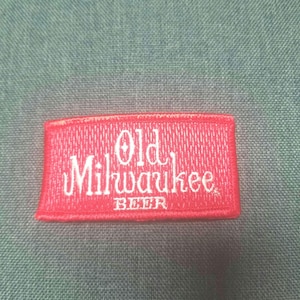 May include: Red fabric patch with white embroidered text that reads "Old Milwaukee Beer".