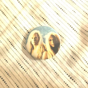 May include: A round button with a picture of two people with long blonde hair. The background is a blue sky with white clouds.