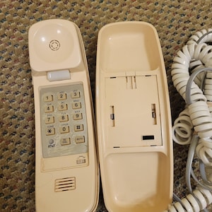 May include: A vintage cream-colored AT&T corded telephone, featuring a keypad with numbers and function buttons. The handset is separated from the base, revealing the interior. A coiled white cord is visible.