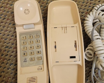 Pode incluir: Um telefone com fio AT&T vintage creme, com um teclado com números e botões de função. O aparelho está separado da base, revelando o interior. Um cabo branco enrolado é visível.