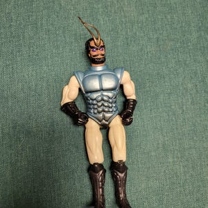 May include: A vintage action figure with a blue and silver torso, tan legs, and black boots and gloves. The figure has a black mustache and beard, purple eyes, and a metallic antenna. The toy is posed with arms at its sides.