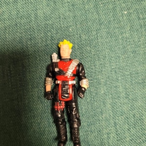 Puede incluir: Una pequeña figura de acción de un hombre con pelo amarillo, vistiendo un atuendo negro y rojo con detalles plateados. La figura tiene un arco y una flecha en la espalda. La figura está sobre una superficie verde.