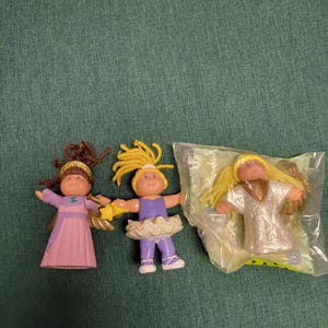 May include: Three Cabbage Patch Kids dolls. One doll wears a pink dress and holds a star. Another doll is dressed in a purple leotard and tutu. The third doll is in a sealed package and wears a white outfit.