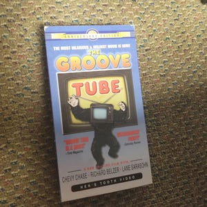 Może przedstawiać: Niebieska kaseta VHS z tytułem "The Groove Tube" w białym kolorze. Kaseta zawiera czarno-białe zdjęcie goryla z telewizorem na głowie. Na kasecie widnieje napis "The Most Hilarious & Wildest Movie Is Here!" Kaseta jest filmem Kena Shapiro z Chevy Chase, Richardem Belzerem i Lane Sarasohn. Kaseta pochodzi z Hen's Tooth Video.