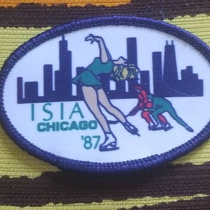 May include: A white circular patch with a blue border. The patch features a silhouette of the Chicago skyline with a figure skater in a green dress and a red and green figure skater in the foreground. The text "ISIA CHICAGO '87" is printed below the skaters.