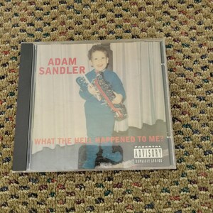 Könnte beinhalten: Eine CD-Hülle mit dem Album "What the Hell Happened to Me?" von Adam Sandler. Das Cover zeigt einen jungen Adam Sandler mit einem Spielzeug. Die CD-Hülle hat einen Warnhinweis für Eltern.