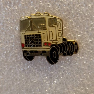May include: A white metal pin of a semi-truck with a black grill and red taillights. The truck has a black cab and a black trailer with black wheels.