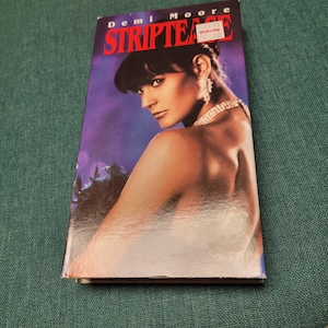 May include: A VHS tape of the movie "Striptease" featuring Demi Moore. The cover has a purple and blue background with Demi Moore's image. The title "Striptease" is in large red letters.