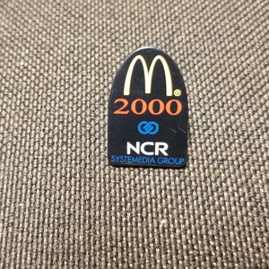 May include: Black and yellow plastic pin with the McDonald's golden arches logo, the year 2000, a blue and orange infinity symbol, and the text "NCR SYSTMEDIA GROUP".