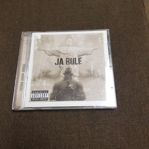 Puede incluir: Una caja de CD con un fondo blanco y una imagen en blanco y negro de un hombre de pie frente a una estatua. El texto "JA RULE" está impreso en letras negras en la caja. La caja también tiene una pegatina de advertencia para padres.
