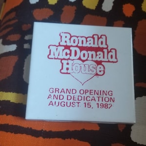 Może przedstawiać: Biała kwadratowa płytka z czerwonym tekstem, który brzmi "Ronald McDonald House" z czerwonym symbolem serca. Tekst poniżej brzmi "Grand Opening and Dedication August 15, 1982."
