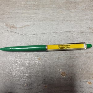 May include: A retractable ballpoint pen featuring a solid green barrel and a yellow mid-section with the black text SCOTTSDALE ARIZONA printed on it. The pen includes a silver-colored metal pocket clip and a green push-button mechanism at the top.