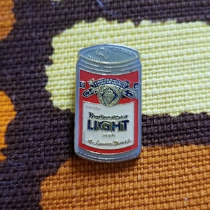 May include: A silver and red Budweiser Light beer can pin. The can features a blue and gold ribbon with the Budweiser logo and the words "Budweiser Light Beer Anheuser-Busch, Inc."