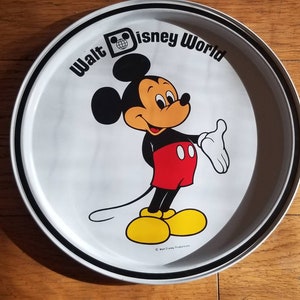 May include: A round, white metal tray featuring Mickey Mouse from Walt Disney World. Mickey is in his classic outfit with red shorts, yellow shoes, and white gloves. The tray has a black and white border and the words "Walt Disney World" are printed around the top.