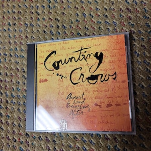 Könnte beinhalten: Eine CD-Hülle für das Album "August and Everything After" von Counting Crows. Das Cover hat einen handschriftlichen Stil mit dem Namen der Band und Songtexten in schwarzer Tinte auf orangefarbenem Hintergrund. Die CD-Hülle ist transparent.