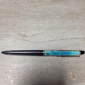 May include: A black ballpoint pen featuring a light blue barrel section with a graphic of the Space Needle. The text on the barrel reads SPACE NEEDLE HEIGHT 605FT SEATTLE U.S.A. The pen includes a silver-coloured metal clip and a black cap.