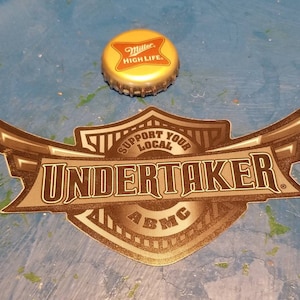 Może przedstawiać: Złota i brązowa naklejka z tekstem "Support Your Local Undertaker" i logo "ABMC".
