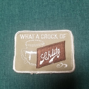 May include: Embroidered patch featuring the text "WHAT A CROCK OF" above a mug illustration and a brown rectangle with "Schlitz" in white script. The patch has a beige background and a white border.