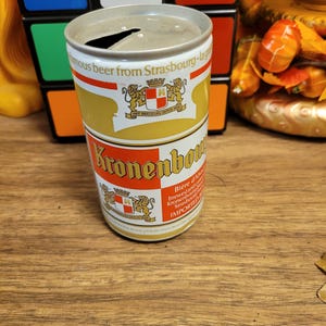 May include: A vintage Kronenbourg beer can with a gold and white label. The label features the words "Kronenbourg" and "famous beer from Strasbourg-lager". The can has a pull-tab opening.