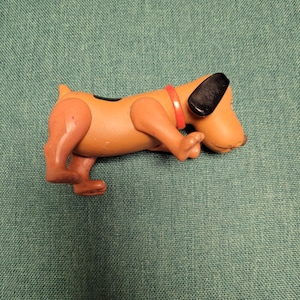 Puede incluir: Un juguete de plástico marrón con forma de perro, con orejas negras y un collar rojo. El perro está en posición de gateo, con las patas delanteras extendidas y las traseras dobladas. El juguete tiene una mancha negra en la espalda.