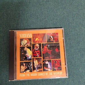 May include: A CD case featuring the band Nirvana. The album cover is orange with multiple images of the band members performing. The text "NIRVANA" is at the top, and "FROM THE MUDDY BANKS OF THE WISHKAH" is at the bottom.