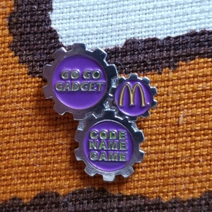 May include: A silver pin with three interlocking gears. The top gear is purple with the text "Go Go Gadget" in yellow. The middle gear is purple with a yellow "M". The bottom gear is purple with the text "Code Name Game" in yellow.