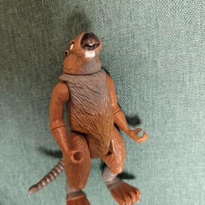 May include: A brown action figure of a rat-like character with a gray vest and gray tail. The figure has articulated joints and is posed on a teal surface. The figure's fur is detailed.