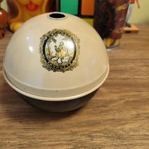 May include: A vintage, cream-colored metal container with a round shape. The container has a dark brown base and a lid with a decorative emblem. The emblem features a woman and sheep. The container is likely for storage.