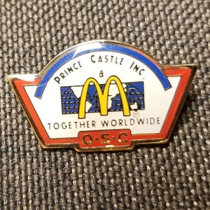 May include: A gold-colored pin with a white background and a red, white, and blue border. The pin features the McDonald's golden arches logo and the text "Prince Castle Inc. & Together Worldwide Q.S.C."