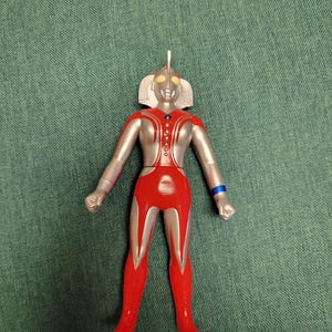 Puede incluir: Figura de acci&oacute;n roja, plateada y azul de un superh&eacute;roe. La figura tiene una cabeza plateada con ojos amarillos y un pecho y piernas rojos. La figura est&aacute; de pie sobre una superficie verde.