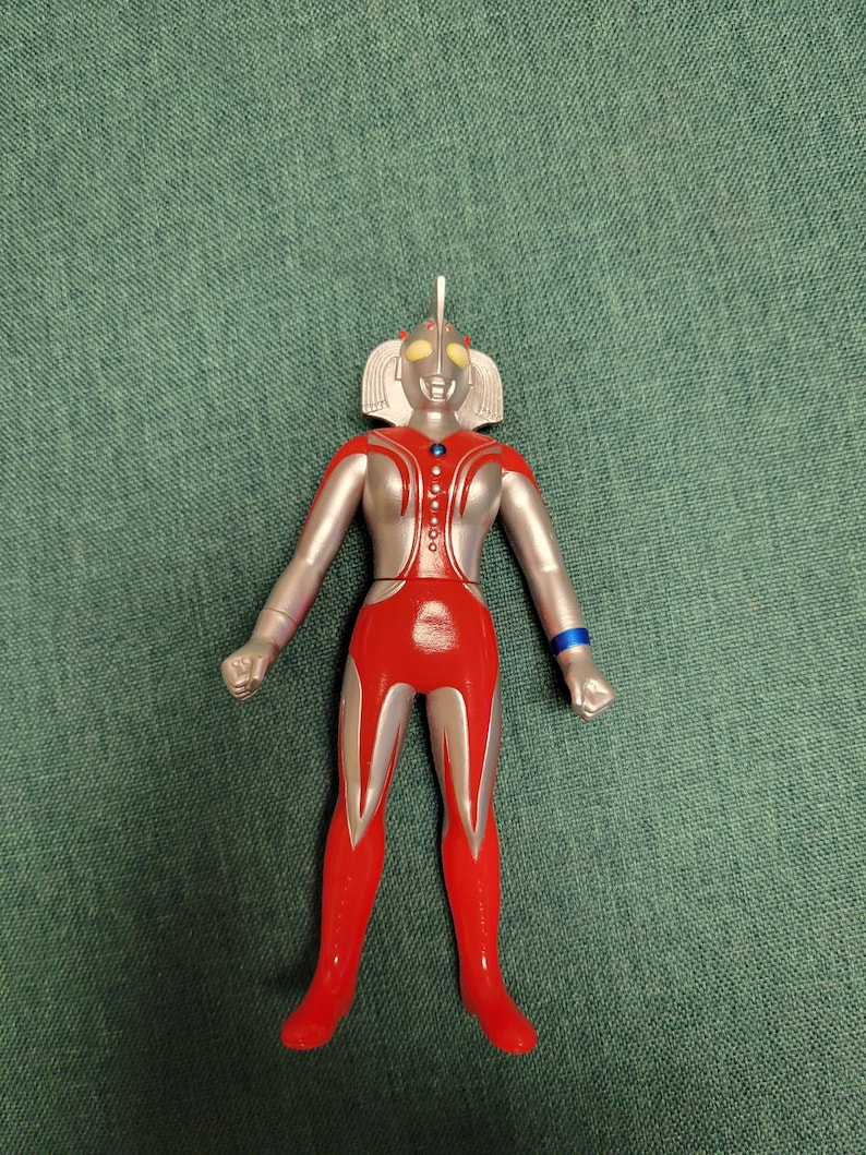 Puede incluir: Figura de acci&oacute;n roja, plateada y azul de un superh&eacute;roe. La figura tiene una cabeza plateada con ojos amarillos y un pecho y piernas rojos. La figura est&aacute; de pie sobre una superficie verde.