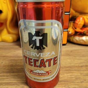Può includere: Una lattina di birra Tecate rossa e argento. La lattina presenta le parole "CERVEZA TECATE" a grandi lettere, con un logo di un'aquila e la lettera "T". La lattina è prodotta in Messico.