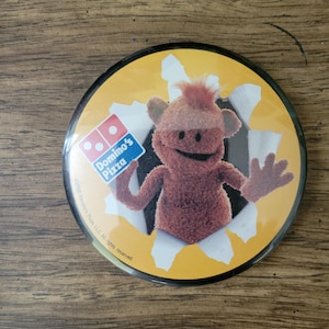 Op de afbeelding: Een ronde button met een stripfiguur die door een gescheurde gele achtergrond komt. Het personage is bruin en harig, met een lachend gezicht. Het Domino's Pizza-logo is zichtbaar.