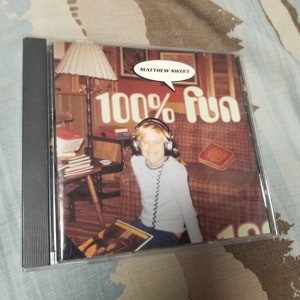 Puede incluir: Una caja de CD negra con una imagen colorida de una persona joven con auriculares y una camisa azul. El texto "MATTHEW SWEET" y "100% fun" están impresos en la caja.