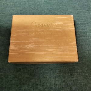 Puede incluir: Una caja rectangular dorada de naipes Caspari, envuelta en plástico transparente. El texto "Caspari PLAYING CARDS" está impreso en la parte superior de la caja. La caja está sobre una superficie verde texturizada.