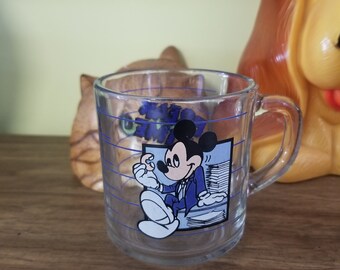 May include: A clear glass mug with a blue and white design featuring Mickey Mouse in a suit. The mug has blue lines around the outside.