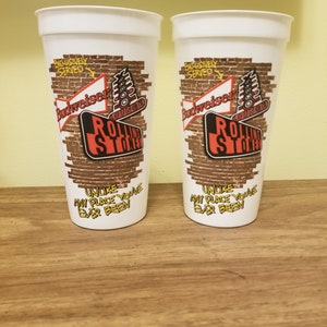May include: Two white plastic cups with a brick wall design. The cups feature the Rolling Stones logo and Budweiser branding. The text on the cups reads "Exclusively Served" and "Unlike any place you've ever been".