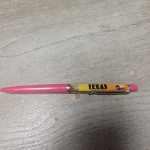 Peut inclure: Un stylo à bille rose doté d'une section de corps jaune avec le texte "TEXAS THE LONE STAR STATE" imprimé en noir, accompagné d'un petit graphique du drapeau de l'État du Texas et d'un bâtiment, fini avec un clip de poche en métal argenté.