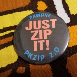Może przedstawiać: Okrągła, czarna przypinka z napisem "PKWARE JUST ZIP IT! PKZIP 2.0" w różowych i niebieskich literach. Przypinka jest umieszczona na wzorzystym tle w kolorach brązu, żółci i pomarańczy.