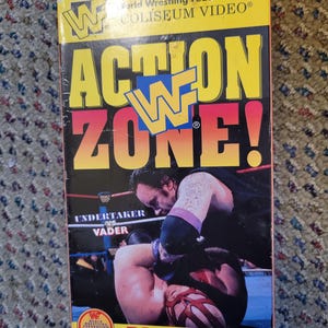 Peut inclure: Une cassette VHS vintage pour "WWF Action Zone!" avec Undertaker contre Vader. La couverture est jaune, rouge et noire avec le logo WWF. Comprend du contenu bonus et des astuces pour les jeux vidéo.