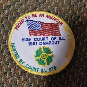 Op de afbeelding: Een witte geborduurde patch met een gele rand. De patch heeft een Amerikaanse vlag, een klavertjevier met de letters "I.O.F." in het midden en de tekst "PROUD TO BE AN AMERICAN AND A FORESTER HIGH COURT OF ILL 1991 CAMPOUT HOSTED BY COURT ILL 878".