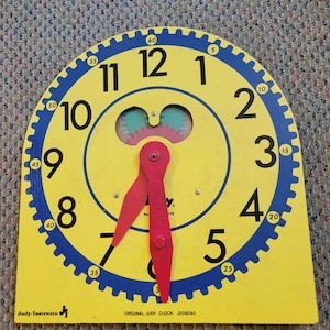 May include: A yellow and blue Judy Clock teaching tool with black numbers 1 through 12 and blue minute markers 5 through 60. It features two red plastic hands, a central gear mechanism, and the text Judy Clock and Original Judy Clock J209040.