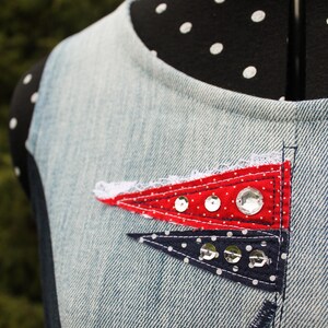 Könnte beinhalten: Jeans-Top mit roten und marineblauen Wimpel-Patches. Der rote Wimpel hat weiße Tupfen und ist mit Pailletten verziert. Der marineblaue Wimpel hat weiße Tupfen und ist ebenfalls mit Pailletten verziert. Das Oberteil hat den Text "JEANS NEW LIFE" auf einem Etikett.