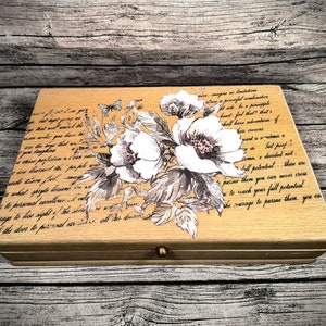 Può includere: Una scatola di legno rettangolare con un motivo floreale e scritte. La scatola ha una finitura marrone chiaro. La parte superiore presenta una grande composizione floreale bianca e grigia con testo scritto. Un piccolo pomello color oro è sul davanti.