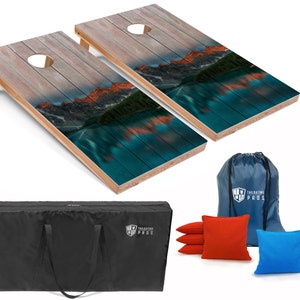 May include: A set of two wooden cornhole boards with a mountain lake scene printed on them. The boards are set up for play with a black carrying bag, red and blue bean bags, and a blue drawstring bag with the words "Tailgating Pros" printed on it.