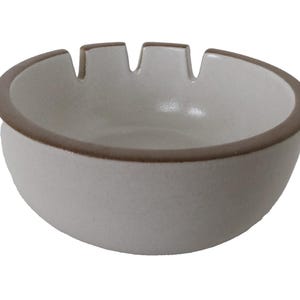 May include: A cream-colored ceramic ashtray with a rounded shape and a brown rim. The ashtray has three notches along the top edge for resting cigarettes. The design is simple and functional.