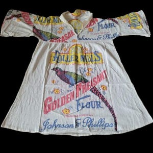 May include: White dress made from repurposed flour sack fabric. The dress features a V-neck and short sleeves. Bold text and graphics include "Golden Pheasant Flour" and a colorful bird illustration. The dress is a unique, vintage garment.