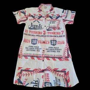 May include: White button-down shirt with a baseball-themed print in red and navy blue. The shirt features baseball bats, balls, and text that reads "Baseball Street N. USA" and "Junk is Turning 7". The shirt has short sleeves and a collared neckline.