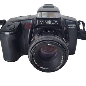 May include: Black Minolta Maxxum 5000i camera with a lens. The camera has a built-in flash and a strap. The lens is labeled "AF 50mm 1:1.7 (22)". The camera body has the Minolta logo.