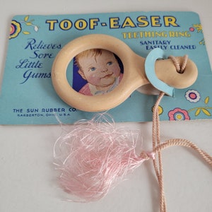 May include: A vintage Toof-Easer teething ring for babies. The ring is made of beige rubber and has a small picture of a baby in the center. The ring is attached to a pink tassel and a beige cord. The packaging says "Toof-Easer Teething Ring, Sanitary Easily Cleaned, Relieves Sore Little Gums, The Sun Rubber Co., Barberton, Ohio U.S.A."